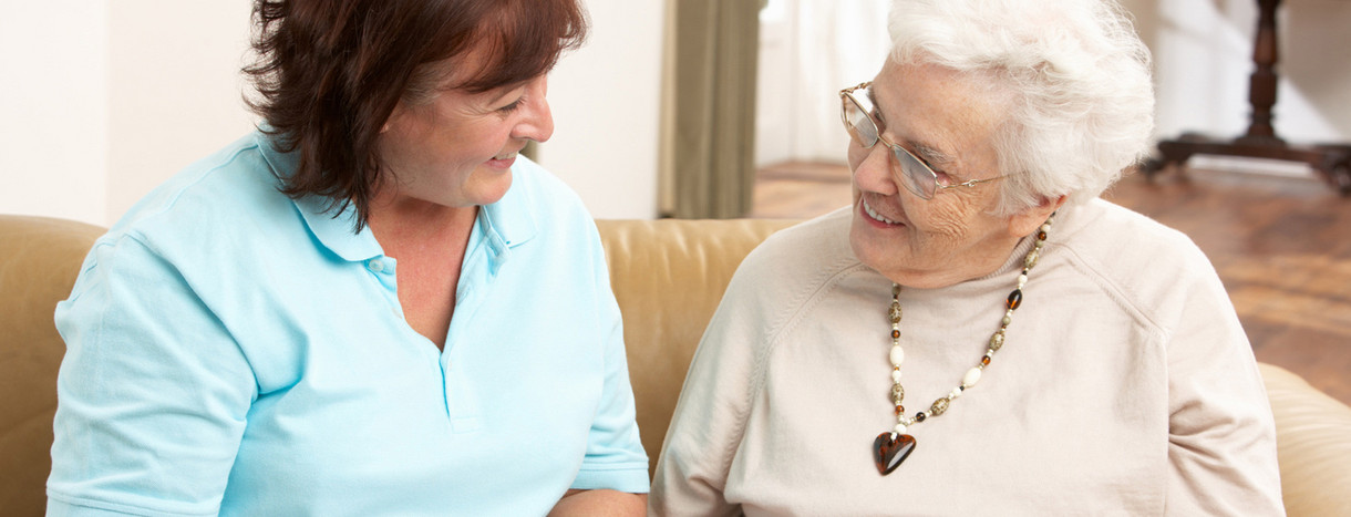 Pflegerin unterhält sich mit älterer Frau in deren Wohnzimmer
