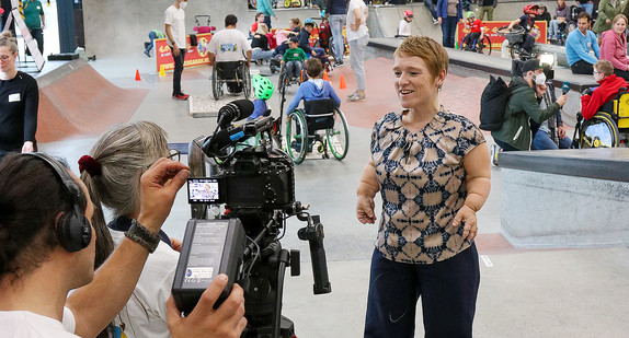 Landes-Behindertenbeauftragte Simone Fischer wird von Reporterin interviewt, im Hintergrund der Parcour des Wheelchair Skills Day.