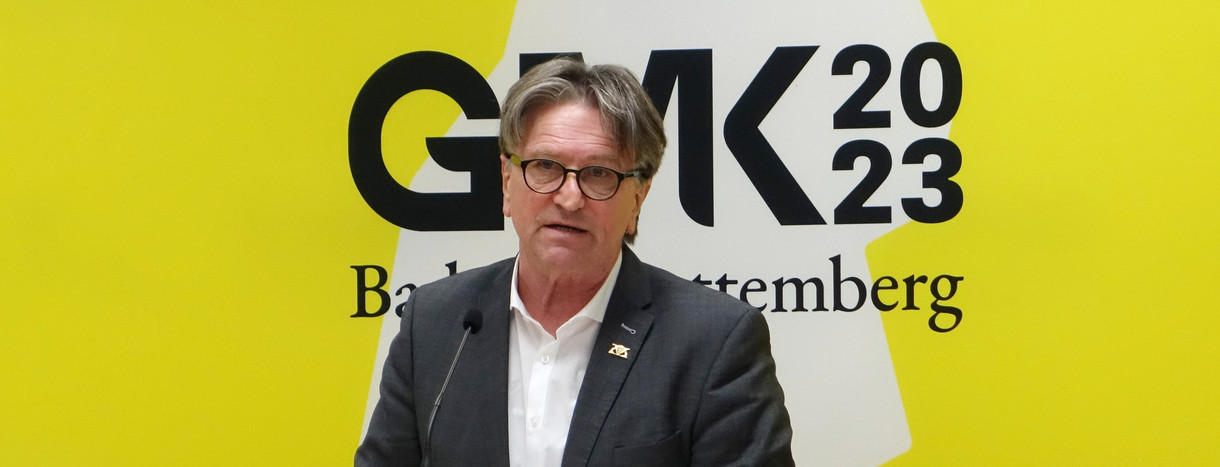 Minister Manne Lucha spricht an Redepult zu Journalisten, im Hintergrund ein großes Banner mit Logo der GMK 2023