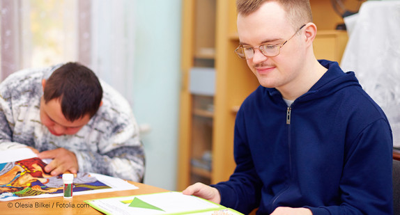 Junger Mann mit Behinderung sitzt an Tisch und liest in einem Lernheft