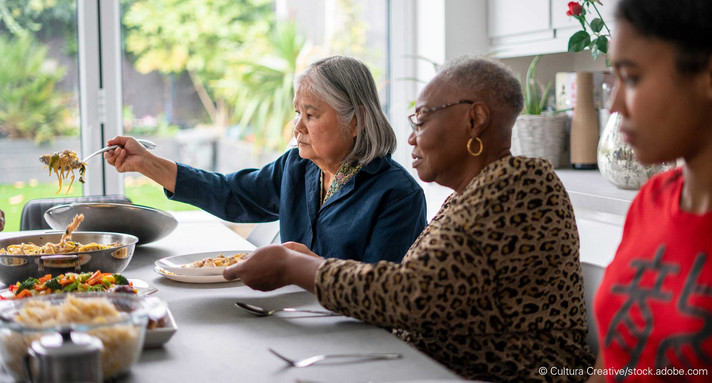 Zwei ältere Frauen sitzen an Esstisch, vor ihnen mit Essen befüllte Schüsseln mit Nudeln und Gemüse.