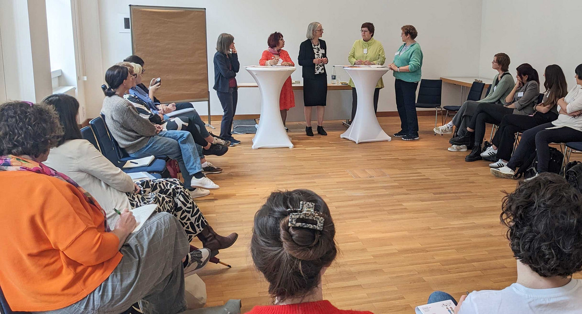 Mehrere Menschen sitzen kreisförmig in einem Raum und hören fünf nebeneinanderstehenden Frauen zu.