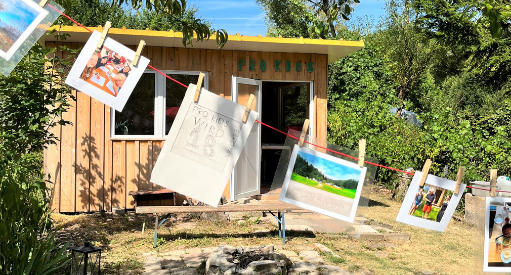 Im Garten sind vor dem Gartenhäuschen an einem Wäscheseil bunte Fotos von Kindern aufgehängt, auf einem Bild steht: Pro Kids wird 18.