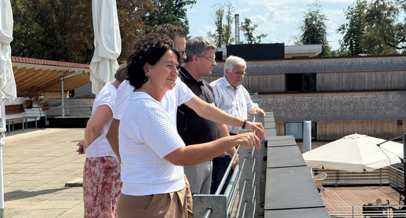 Frau Staatssekretärin Petra Krebs steht auf einem Balkon des Altenhilfezentrums und lässt sich von Verantwortlichen den Aufbau der Anlage erklären.