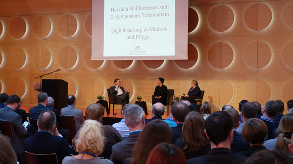 Gesundheitsminister Manne Lucha, Moderatorin Britta March und Wissenschaftsministerin Theresia Bauer auf der Bühne im Hospitalhof Stuttgart beim zweiten Symposium „Telemedizin - Digitalisierung in Medizin und Pflege“