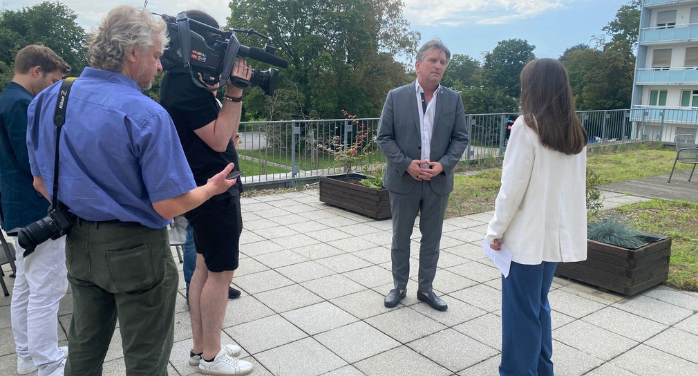 Minister Lucha beantwortet auf der Klinikterrasse in Mannheim Fragen der anwesenden Medienvertreterinnen und Medienvertreter. In der linken Bildhälfte sieht man einen Kameramann, der den Minister filmt.