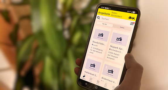 Eine Person hält ein Smartphone in der Hand. Auf dem Display sieht man die Startseite der Ehrenamtskarten-App für Baden-Württemberg. 