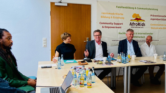 Minister Lucha sitzt mit Mitgliedern des Vereins Afrokids International Stuttgart zusammen. Im Hintergrund hängt ein großes Banner mit den Vereinsthemen.