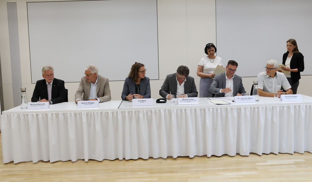 Minister Manne Lucha und Vertreter der vier Modellstandorte sitzen an einem langen Tisch vor Publikum.