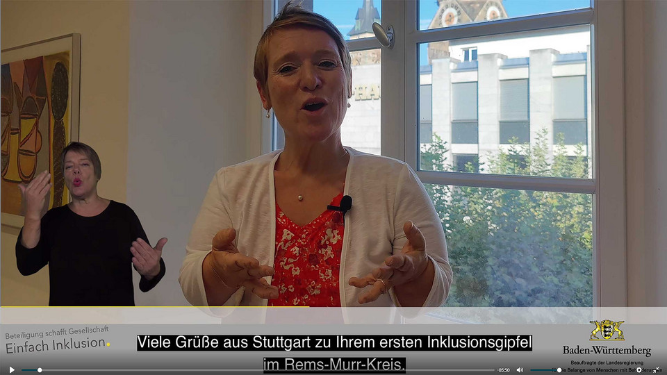 Screenshot aus Video-Grußwort von Simone Fischer, stehend vor einem Fenster mit Ausblick auf Bäume.