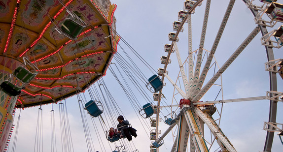 Zwei Kinder die in einem Riesenkarusell sitzen und es ist ein Riesenrad zu sehen.