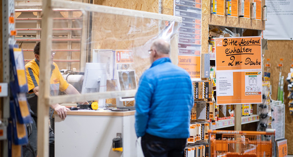 Ein Mann steht in einem Baumarkt am Zuschnitt für Holz in vor einer Plexiglasscheibe.