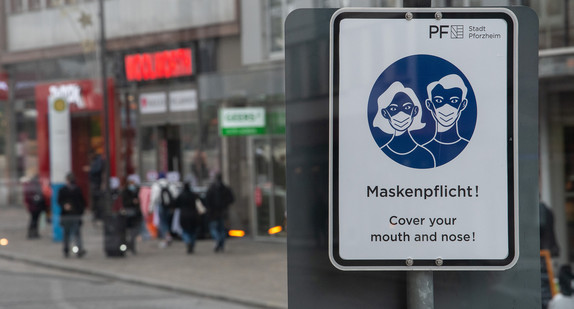 Ein Schild weist in einer Innenstadt auf die Maskenpflicht hin.