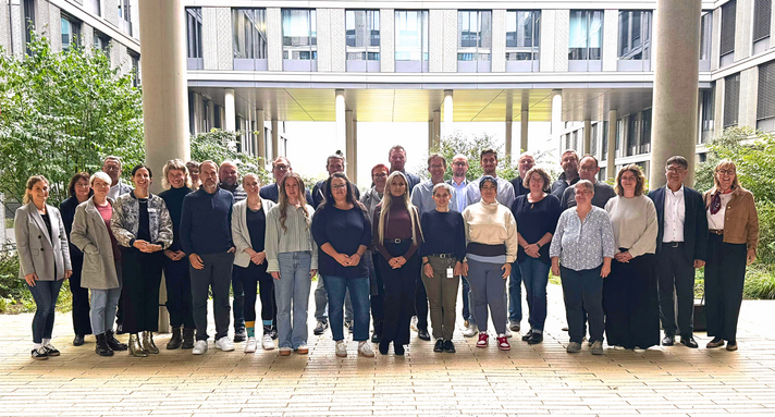 250925_Housing_First_2434x1310 - 1 Die Verantwortlichen für die Pilotprojekte der Initiative Housing First haben sich in einem begrünten Innenhof zum Gruppenfoto aufgestellt.