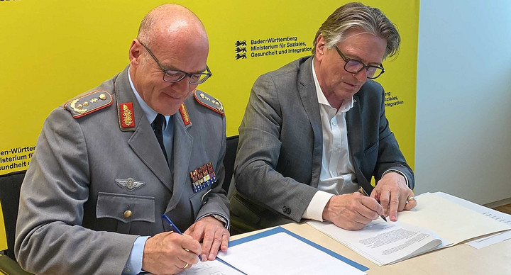 Kommandeur der Sanitätsakademie der Bundeswehr, Generalstabsarzt Dr. Hans-Ulrich Holtherm und Gesundheitsminister Manne Lucha sitzen nebeneinander und unterzeichnen jeweils ein Dokument.