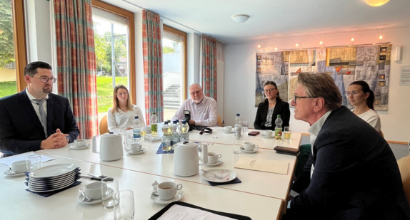 Gesundheitsminister Manne Lucha lässt sich in einem Besprechungsraum der Gesundheitspraxis Wilhelmsdorf das Konzept der innovativen Praxis erklären. Insgesamt sechs Personen sitzen um den Besprechungstisch. Im Hintergrund sieht man ein großformatiges, abstraktes Bild.