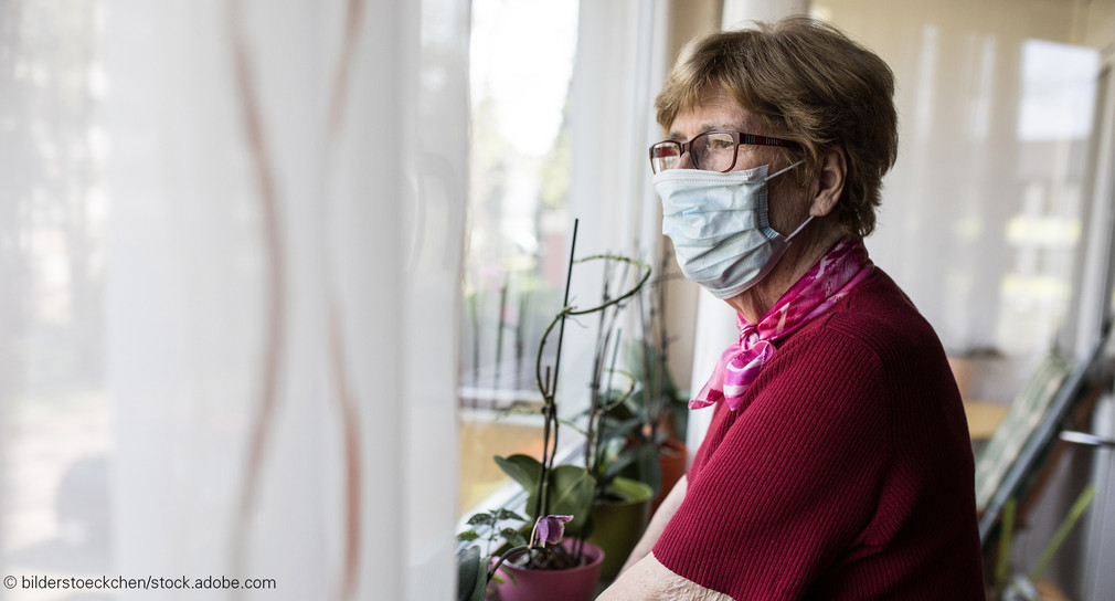 Ältere Frau mit OP-Maske schaut aus Wohnungsfenster