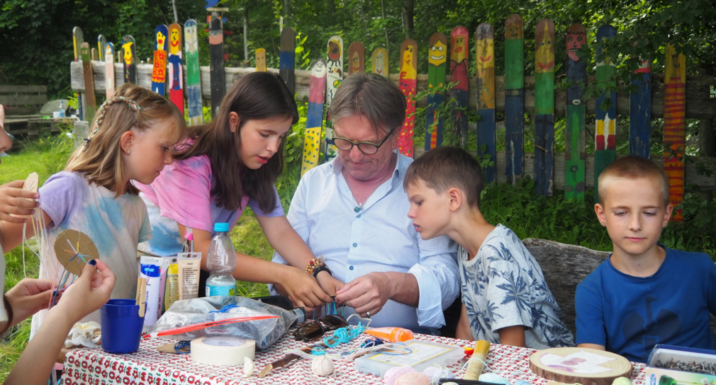 Der Minister bastelt mit Kindern aus der Ferienfreizeit. Sie sitzen im Freien an einem Tisch.