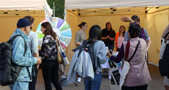 Menschengruppe steht an einem Infostand vor einem Glücksrad