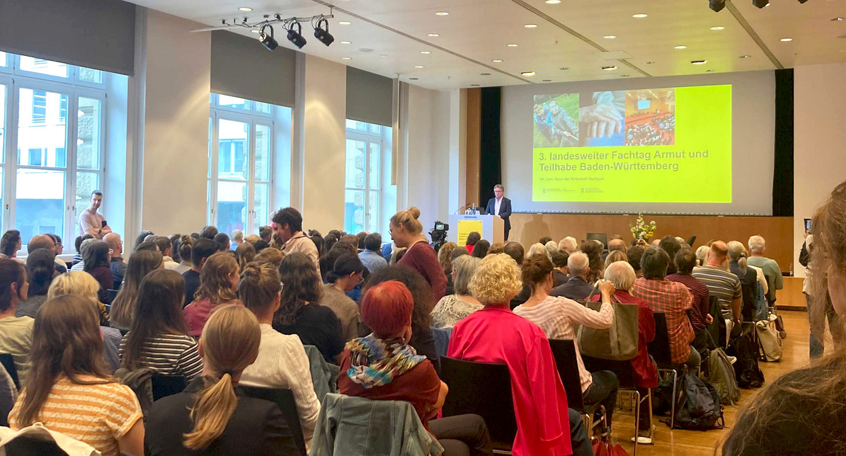 Minister Manne Lucha steht auf Bühne vor Publikum in einem Veranstaltungssaal, im Hintergrund eine große Leinwand mit dem Titel der Veranstaltung: 3. landesweiter Fachtag Armut und Teilhabe Baden-Württemberg.