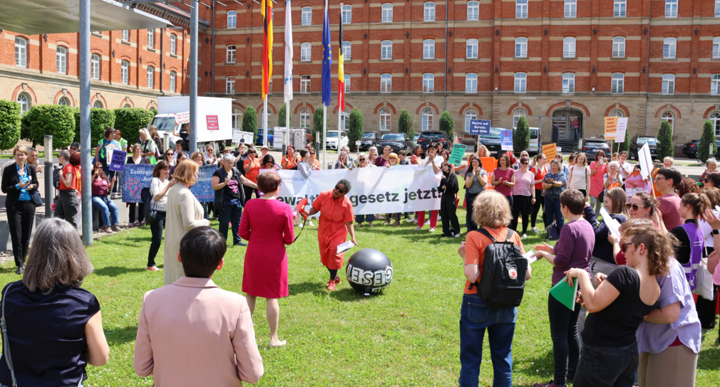 Demonstrierende von den autonomen Frauenhäusern nutzen die Chance zum Gespräch mit den Ministierinnen. Der Austausch findet bei Sonnenschein im Freien statt. Viele Frauen sind mit Plakaten und Bannern erschienen.