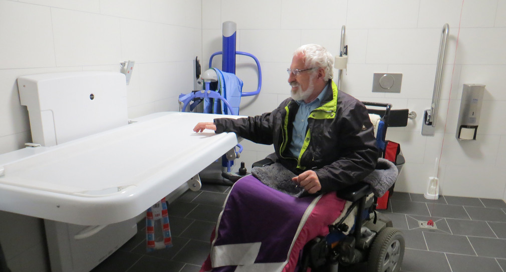Mann im Rollstuhl testet die höhenverstellbare Wandklappliege für Erwachsene in einer „Toilette für alle“