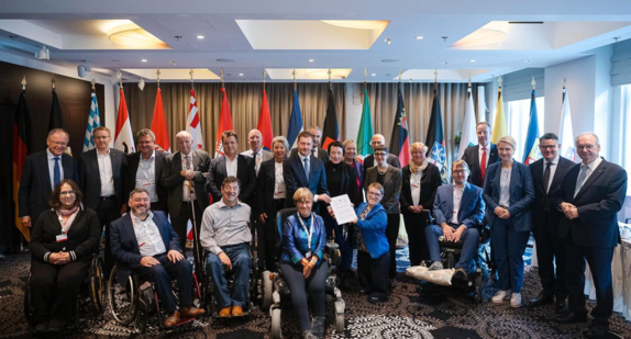 Mitglieder der Jahreskonferenz der Ministerpräsidentinnen und Ministerpräsidenten und die Beauftragten der Länder und des Bundes für die Belange von Menschen mit Behinderungen beim Gruppenbild in Leipzig. Im Hintergrund sieht man die Landesflaggen aller Bundesländer.