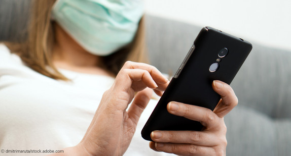 Frau mit OP-Maske sitzt mit Smartphone auf Sofa