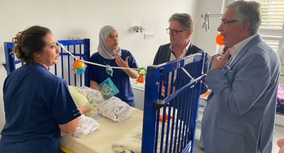 Minister Manne Lucha steht an einem Pflegebett für Kinder. Zwei Pflegeschülerinnen erklären ihm, wie kleine Kinder richtig betreut werden.
