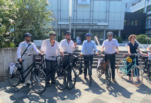 Minister Lucha mit weiteren Teilnehmerinnen und Teilnehmern beim Besuchstermin in der Klinik Diako Mannheim vor dem Gebäude. Alle sind mit Fahrrad und Helm ausgestattet.