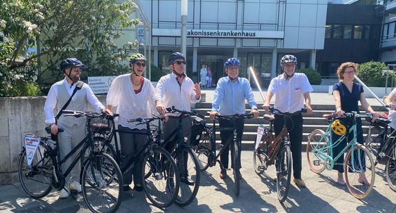Minister Lucha mit weiteren Teilnehmerinnen und Teilnehmern beim Besuchstermin in der Klinik Diako Mannheim vor dem Gebäude. Alle sind mit Fahrrad und Helm ausgestattet.