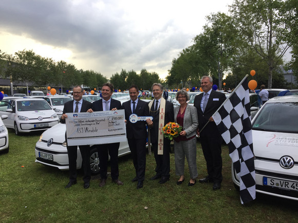 Minister Manne Lucha steht zusammen mit Jürgen Rehm (Vorstand des Gewinnspielvereins Baden-Württemberg e. V.) und Dr. Roman Glaser (Präsident des Baden-Württembergischen Genossenschaftsverbands) vor mehreren Autos und übergibt symbolischen Scheck und Fahrzeugschlüssel