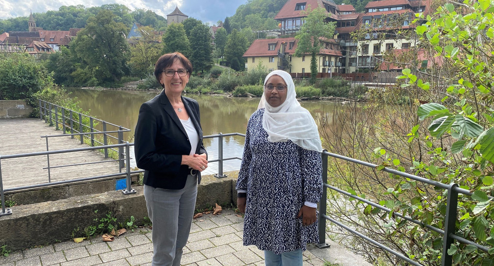Staatssekretärin Dr. Ute Leidig und Frau Hala Elamin, die interkulturellen Promotorin des Vereins Freundeskreis Afrika e. V. stehen für ein Gruppenbild auf der Terrasse. Im Hintergrund sieht man ein idyllisches Flusspanorama.
