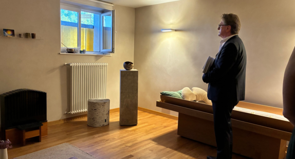 Minister Manne Lucha steht in einem sehr harmonisch gestalteten Abschiedsraum, der durch gedämpftes Licht ausgeleuchtet ist. Ein Fenster ist geöffnet. Vor dem Fenster steht eine Stele mit einer kunstvollen Schale, daneben ein schlichter Hocker. Die schlicht aber sehr geschmackvoll gestaltete Liege für die Verstorbenen ist leer. Der gesamte Raum wirkt warm und würdevoll.