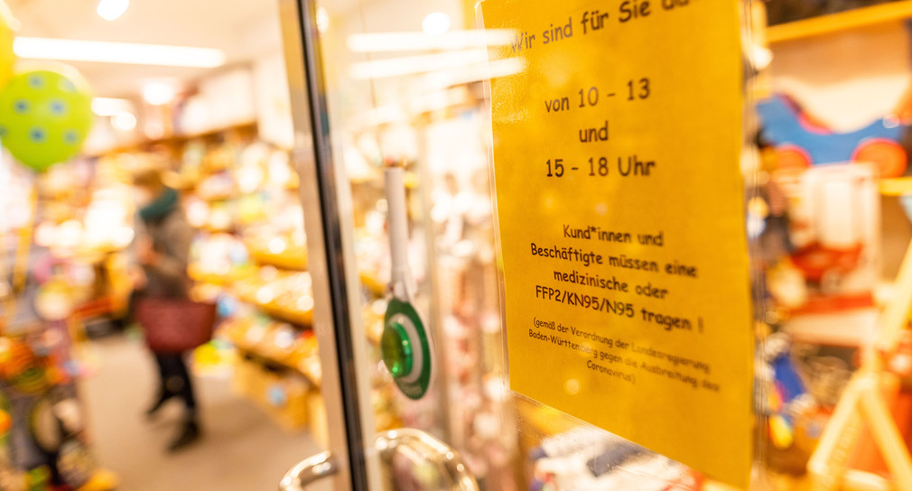 Ein Aushang an der Tür eines Spielzeugladens mit Informationen auf Öffnungszeiten, im Hintergrund steht eine Kundin vor einem Regal.