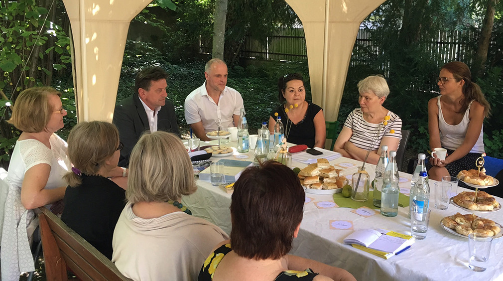 Minister Manne Lucha und Landtagsabgeordneter Abgeordneter Thomas Poreski sitzen mit Vertreterinnen des Mütterforums Baden-Württemberg an Tisch im Freien