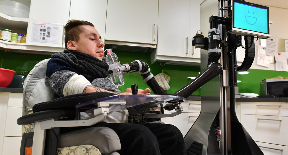 Rollstuhlfahrer bedient mit einer Mundsteuerung einen Roboter, der ihm eine Flasche Wasser reichen soll.