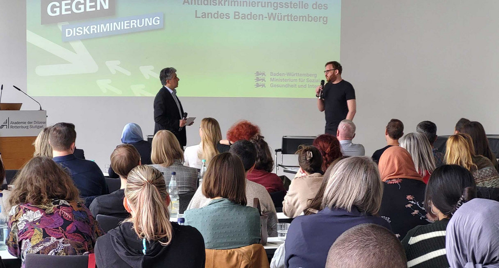Zwei Männer stehen vor Publikum in einem Veranstaltungssal, im Hintergrund eine Leinwand mit Logo der Antidiskriminierungsstelle des Landes Baden-Württemberg