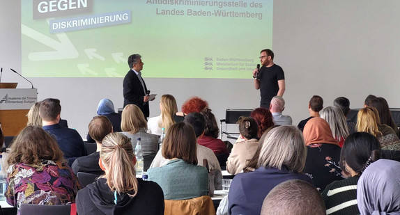 Zwei Männer stehen vor Publikum in einem Veranstaltungssal, im Hintergrund eine Leinwand mit Logo der Antidiskriminierungsstelle des Landes Baden-Württemberg