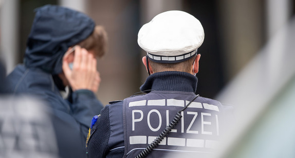 Ein Polizist steht in der Innenstadt vor einem jungen Mann, der seinen Mund-Nasenschutz anlegt, nachdem er von dem Polizisten darauf hingewiesen worden ist.