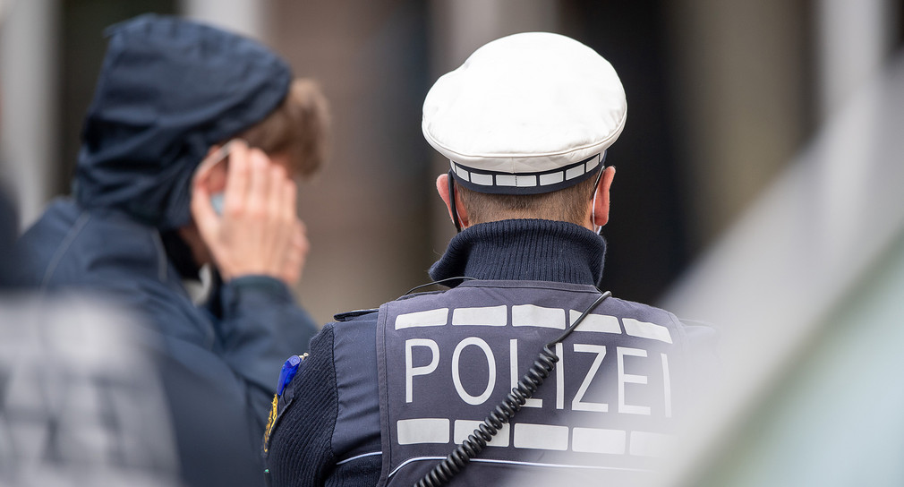 Ein Polizist steht in der Innenstadt vor einem jungen Mann, der seinen Mund-Nasenschutz anlegt, nachdem er von dem Polizisten darauf hingewiesen worden ist.