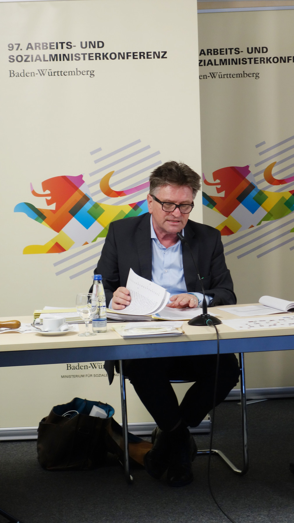 Sozial- und Integrationsminister Manne Lucha sitzt am einem Konferenztisch, hinter ihm steht ein Aufsteller der ASMK 2020