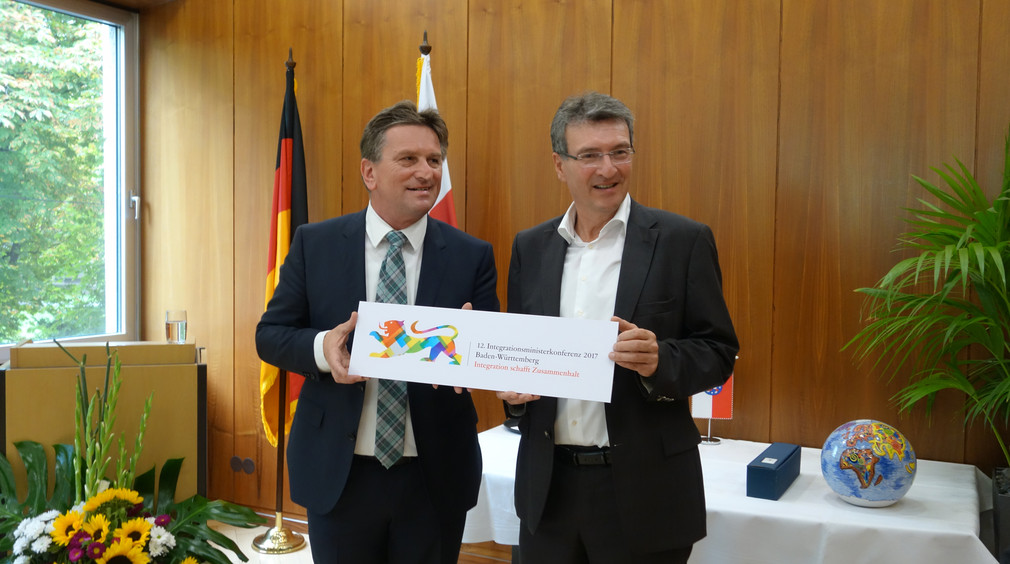 Sozial- und Integrationsminister Manne Lucha und Thüringens Migrationsminister Dieter Lauinger halten Platte mit aufgedrucktem Logo der IntMK 2017
