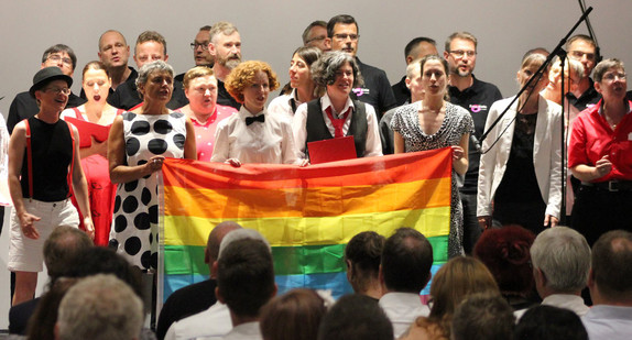 Singende Menschen auf einer Bühne halten zusammen eine Regenbogenflagge
