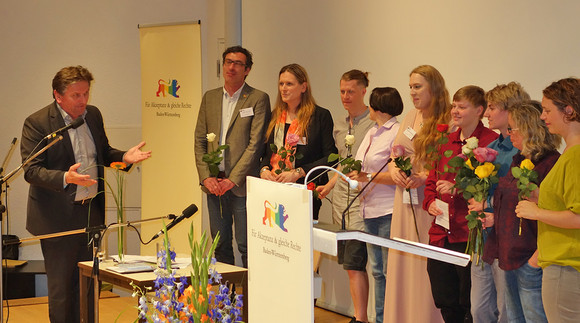 Minister Lucha spricht auf Bühne zu Mathias Falk (Rosa Hilfe Freiburg e.V.), Tamara Kailuweit (Netzwerk LSBTTIQ Baden-Württemberg), Christoph Leder (Projekt „Vielfalt fo(e)rdert!), Margret Göth (PLUS e.V.) und Isabelle Hlawatsch (Projekt „TeenGender“) 