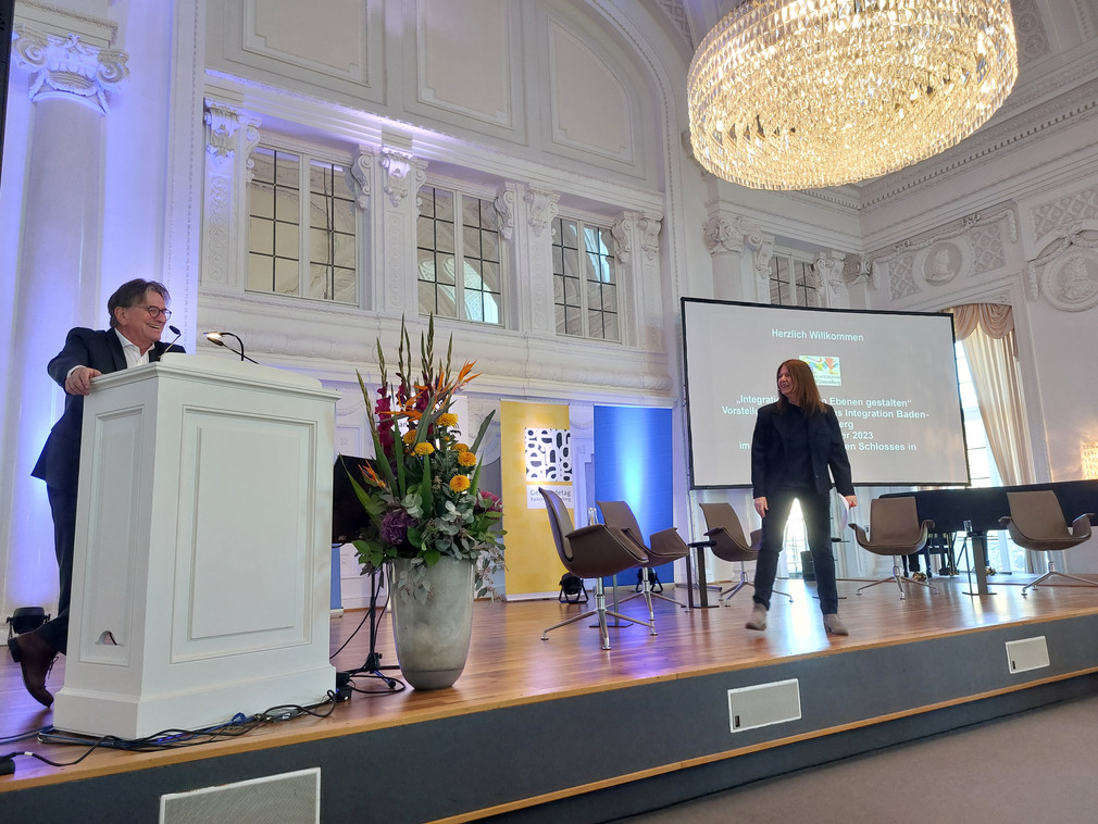 Minister Manne Lucha und Moderatorin Susanne Babila (SWR) stehen auf einer Bühne vor Publikum.