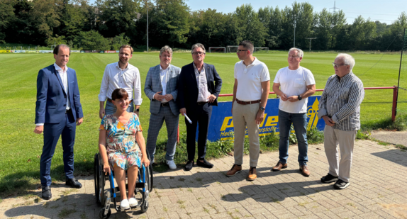 Minister Manne Lucha und Nora Welsch, die Landes-Behindertenbeauftragte, haben sich mit Verantwortlichen des Bauvorhabens zum Gruppenbild versammelt. Frau Welsch nutzt einen Rollstuhl. Da der Bau noch nicht begonnen wurde, sieht man im Hintergrund nur grüne Wiese.