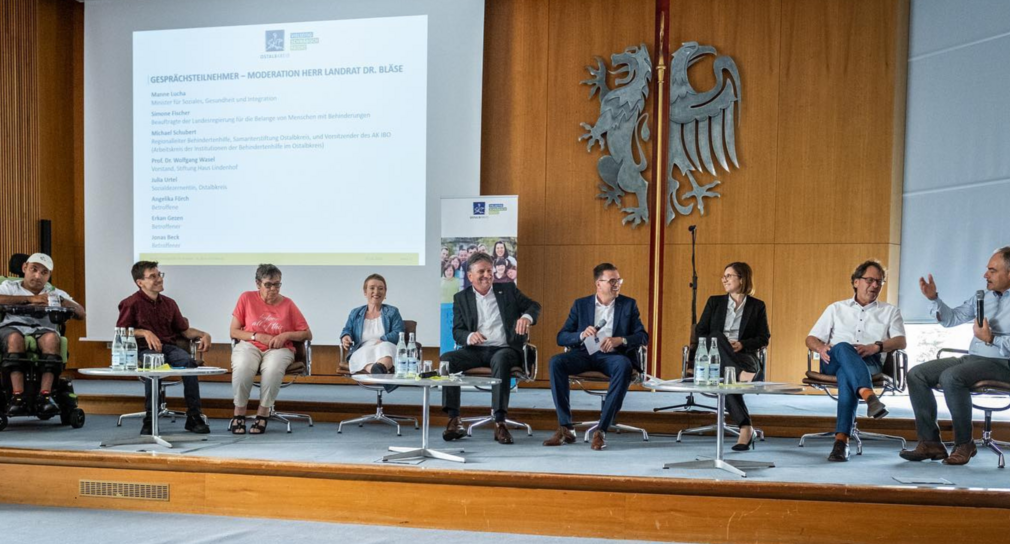 Das Bild zeigt auf der Bühne eine Podiumsrunde (sitzend) von links nach rechts: Erkan Gezen, Jonas Beck, Angelika Förch (für die Perspektive der Menschen mit Behinderungen), Landes-Beauftragte Simone Fischer, Minister Manne Lucha, Landrat Dr. Joachim Bläse, Julia Urtel (Sozialdezernentin), Michael Schubert (Sprecher Trägerforum EGH des Oatalbkreises), Prof. Dr. Wasel (Stiftung Haus Lindenhof)
