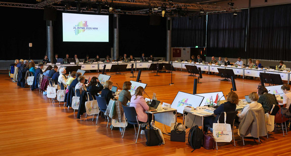 Teilnehmende der Integrationsministerkonferenz 2026 sitzen in großer Runde an Konferenztisch, das Logo der IntMK ist auf eine große Leinwand projiziert.