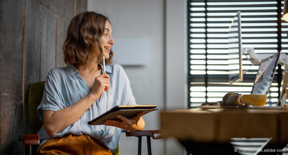 Junge Frau sitzt mit Tablet vor einem Tisch, auf dem ein Laptop steht.
